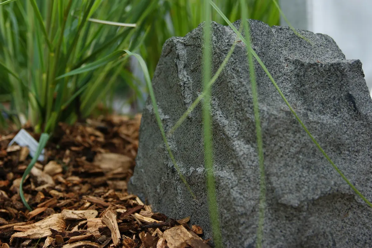 Sonance Rock 63 in Steinform zwischen Gräsern auf Rindenmulch